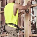 Construction worker in a yellow vest measuring a wooden frame with a tape measure.