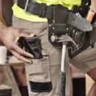 Person in work gear with tool belt, holding phone, hammer and screwdriver visible.