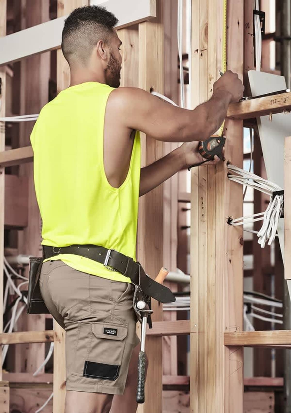 Builder in yellow tank top measures timber framework with a tape measure.