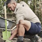 Person gardening, kneeling and using a green shovel, wearing boots and shorts.