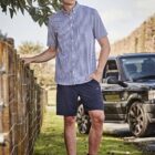 Man in checkered shirt and shorts stands by fence, with an SUV parked behind.