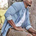 Man in blue shirt and shorts sitting on a rock by a tree, looking into the distance.