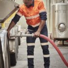 Worker in safety gear connecting a hose to a tank in an industrial setting.