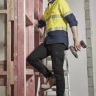 Person in workwear standing on a ladder with a drill, near wooden framework.