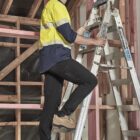 A construction worker climbs a ladder inside a wooden framed building.