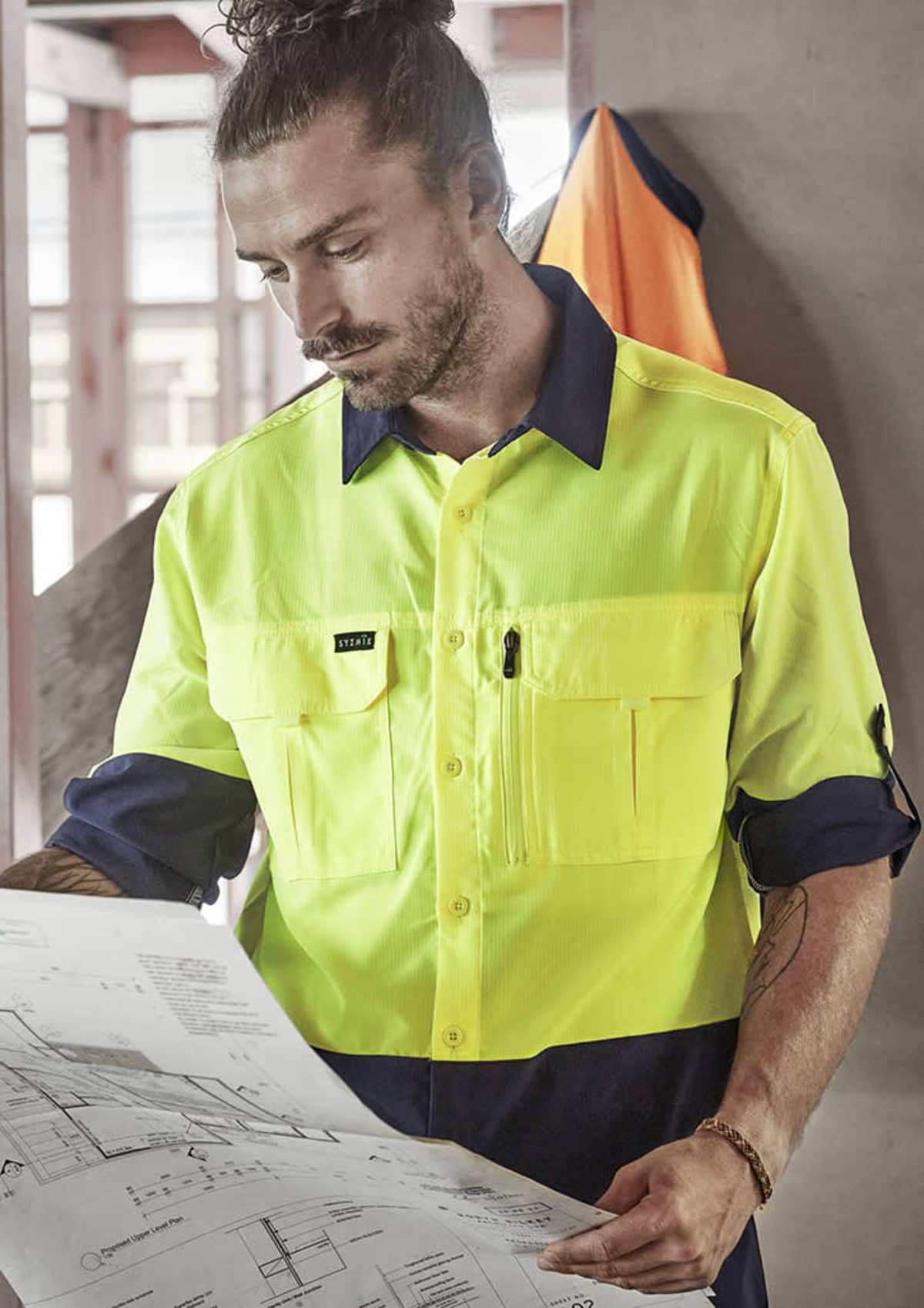 Worker studying blueprints in a construction setting, wearing a yellow safety shirt.