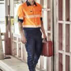 Person in an orange work shirt walks in a construction area, holding a red container.