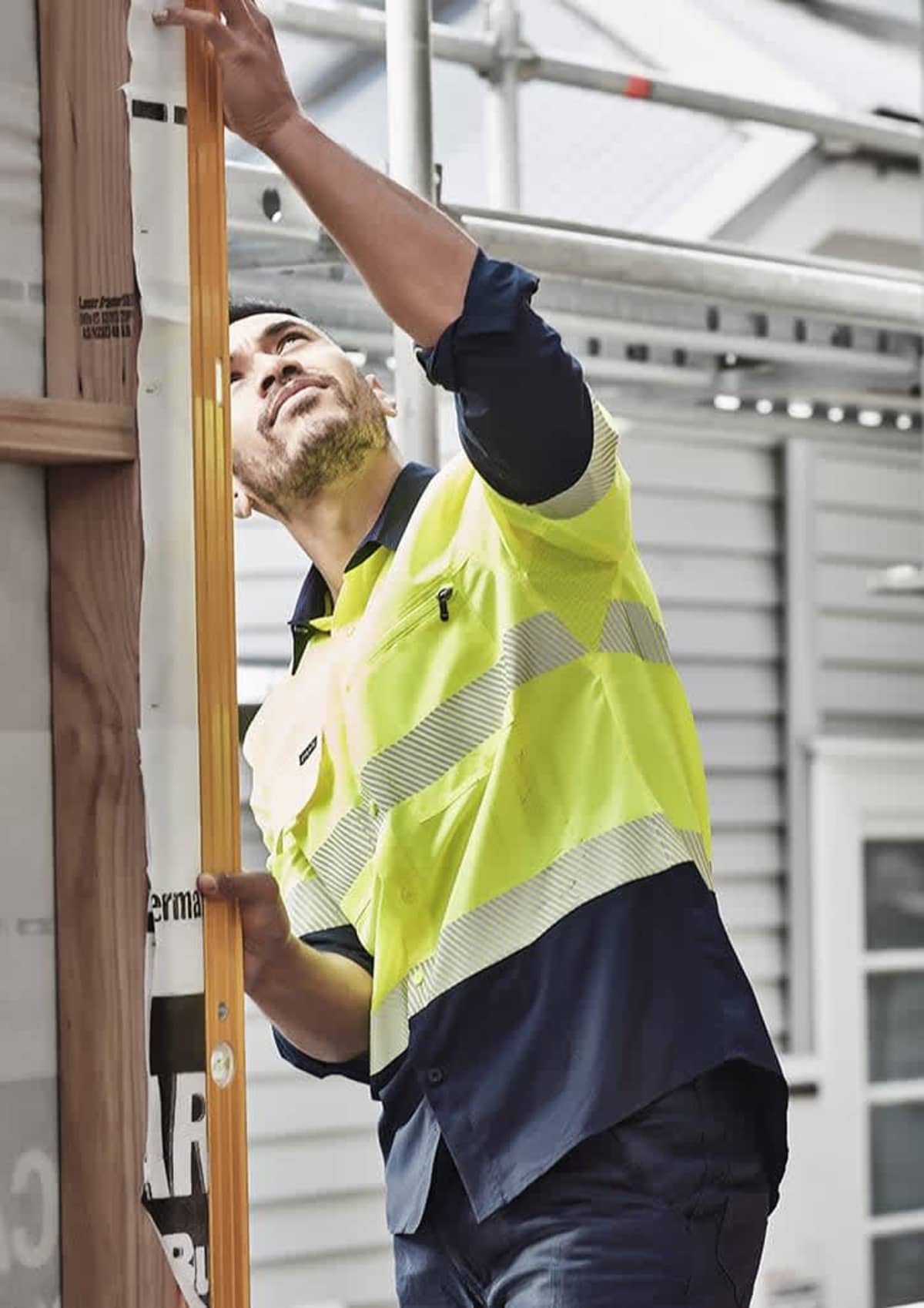 Worker in a reflective vest measures a wooden beam beside a building under construction.