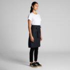 Woman in white t-shirt, black apron, black pants, and sneakers standing against gray background.