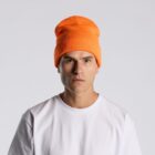 Man in white shirt and orange beanie hat looks at camera against plain light background.