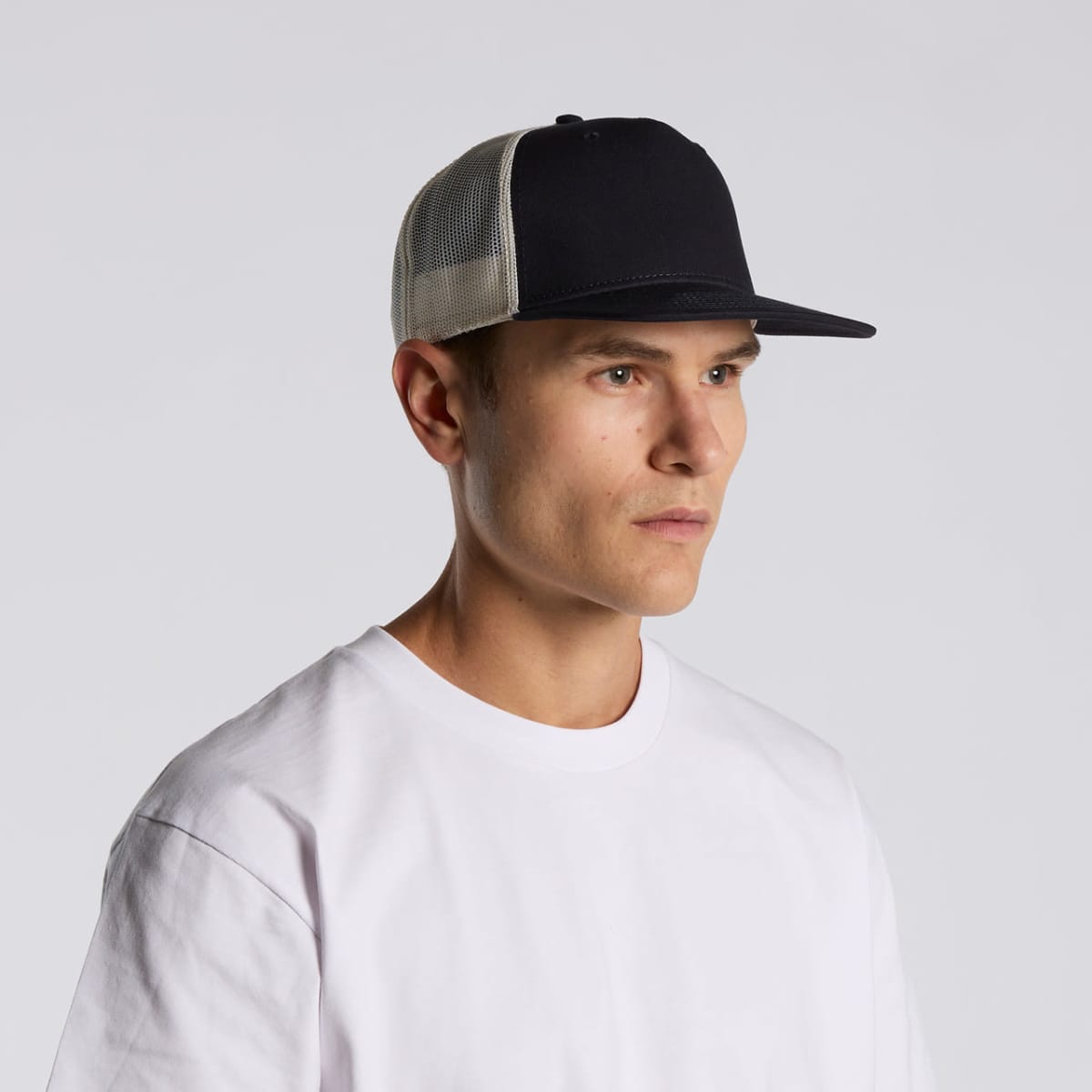 Man in a white t-shirt and black trucker hat looking to the side against a gray background.