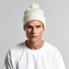 Man in white beanie with pom-pom and white t-shirt against a plain background.