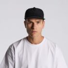 Man in a black cap and white t-shirt staring seriously at the camera against a gray background.