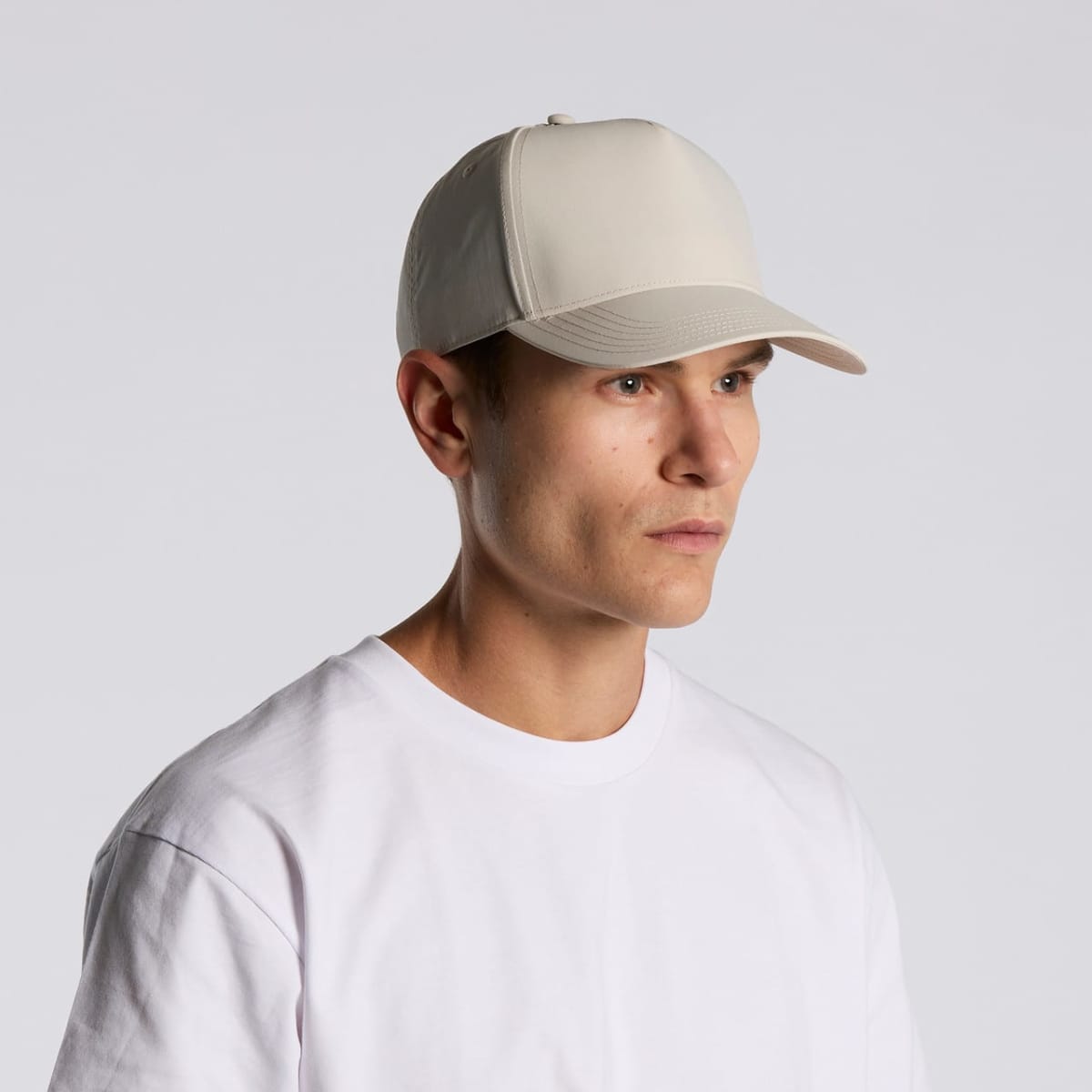 Man in a white T-shirt and beige baseball cap looking to the side against a gray background.