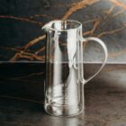 A clear glass pitcher with a handle on a dark marble countertop and background.