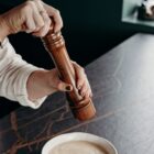 Hands grinding pepper over a bowl of creamy soup on a dark countertop.