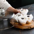 A hand lifts a glass dome to reveal cupcakes topped with cream and berries on a wooden tray.