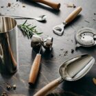 Bar tools with wooden handles and a metal shaker on a dark stone surface with rosemary.