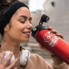 Smiling woman with headphones drinks from a red water bottle outdoors.