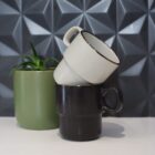 Two stacked mugs beside a green potted plant on a white surface with a geometric background.