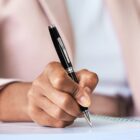 Hand holding a black pen, writing on paper with a spiral notebook.