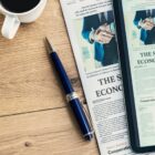 Tablet, newspaper, pen, and coffee cup on a wooden desk, displaying economic news.