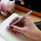 Hand with red nails holds a silver pen over a lined notebook, with pink flowers nearby.
