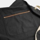 Black apron with a front pocket and a tan leather trim, lying flat on a light surface.