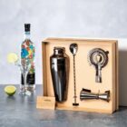 Cocktail shaker set with tools in a box, bottle, glasses, and limes on a table.