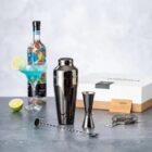Cocktail shaker set with a jigger, strainer, and drink beside a liquor bottle and lime.