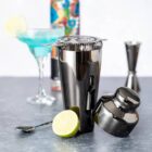 Metal cocktail shaker, jigger, spoon, lime, and blue cocktail glass on a gray surface.