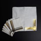White napkins with shiny gold stripe patterns in one corner, displayed on a black background.