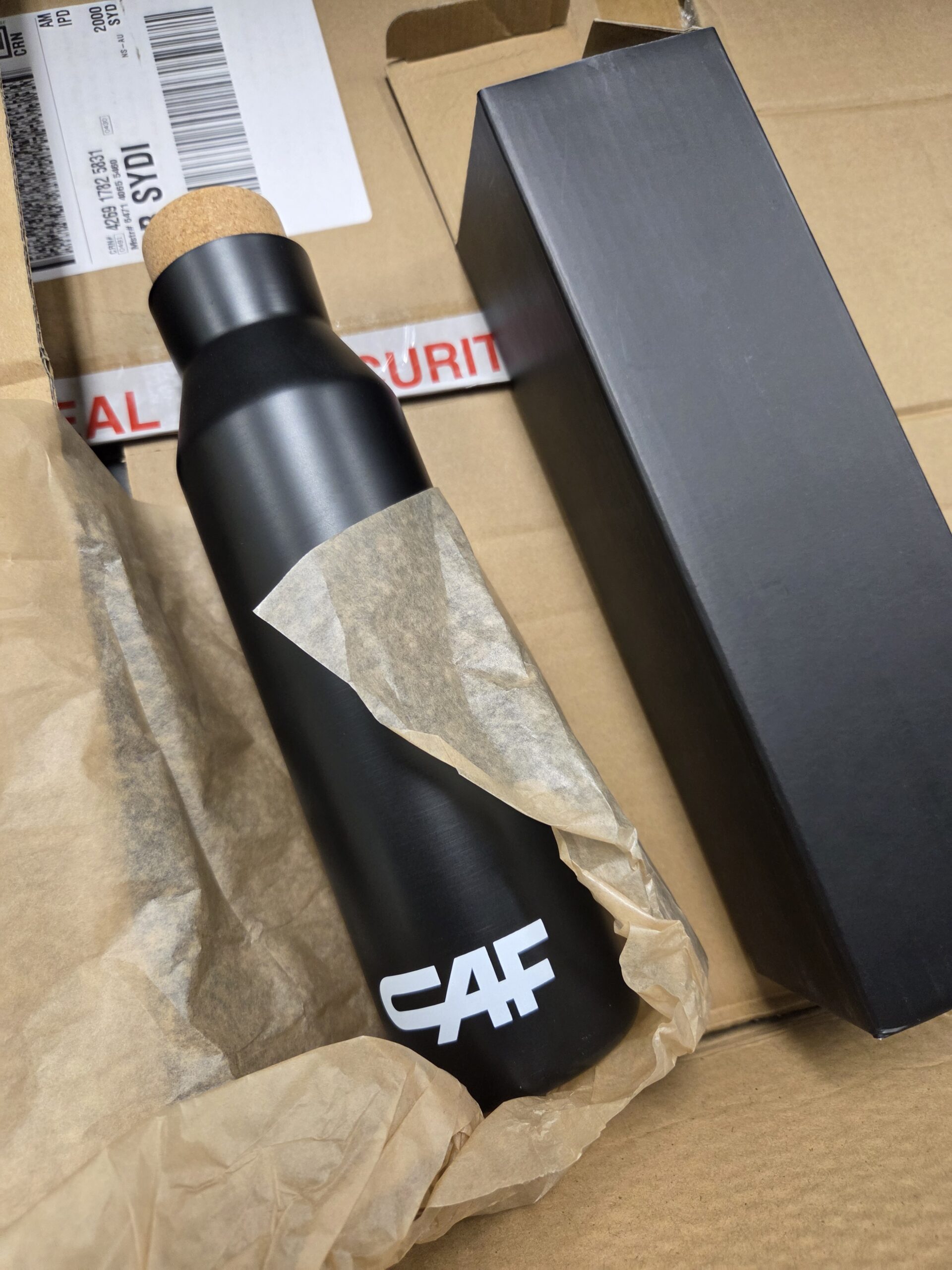 A black metal bottle with cork cover and a black packaging
