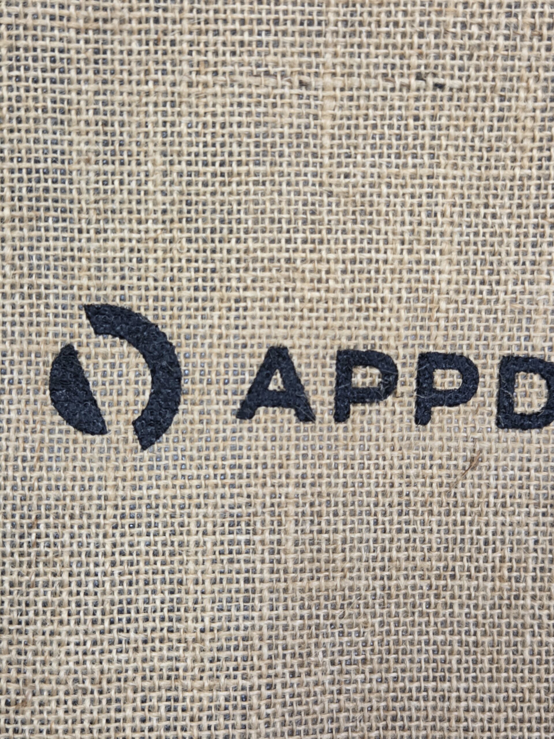 Black text APPD and a circular logo on a burlap textured cotton tote bag background.