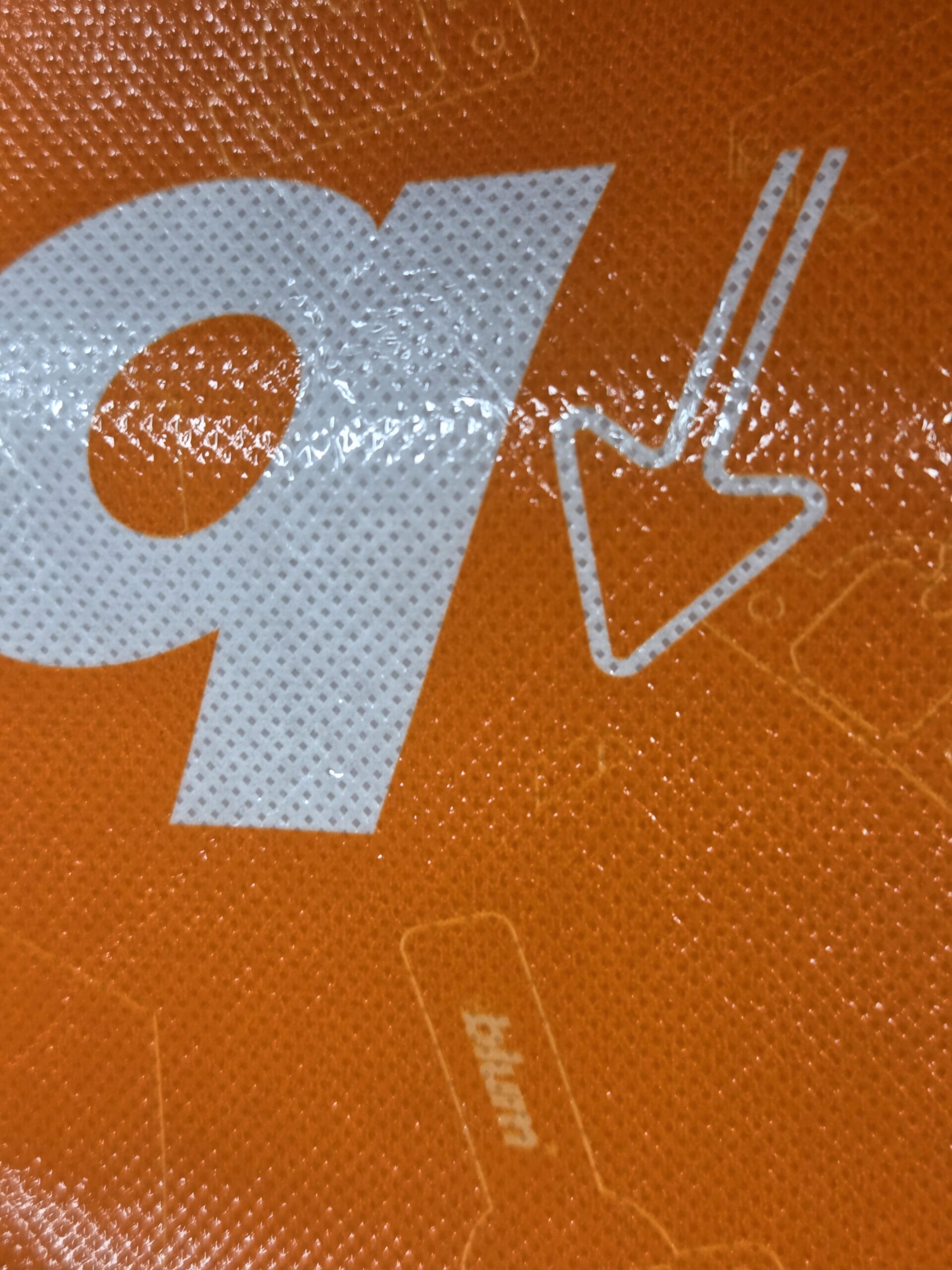 Close-up of a white lowercase q and downward arrow on an orange cotton tote bag surface.