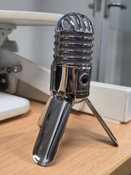 A shiny, vintage-style podcast microphone on a small tripod sits on a wooden desk.