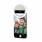 Smartphone with selfie ring light shows two people smiling outdoors on its screen.