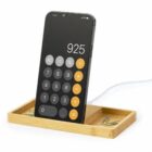 A smartphone displaying a calculator app rests on a wooden charging stand with paperclips.