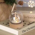 A glass snow globe with wooden trees sits on a book titled The Christmas.