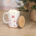 White mug with red snowflakes, filled with marshmallows, beside a wooden lid on a table.