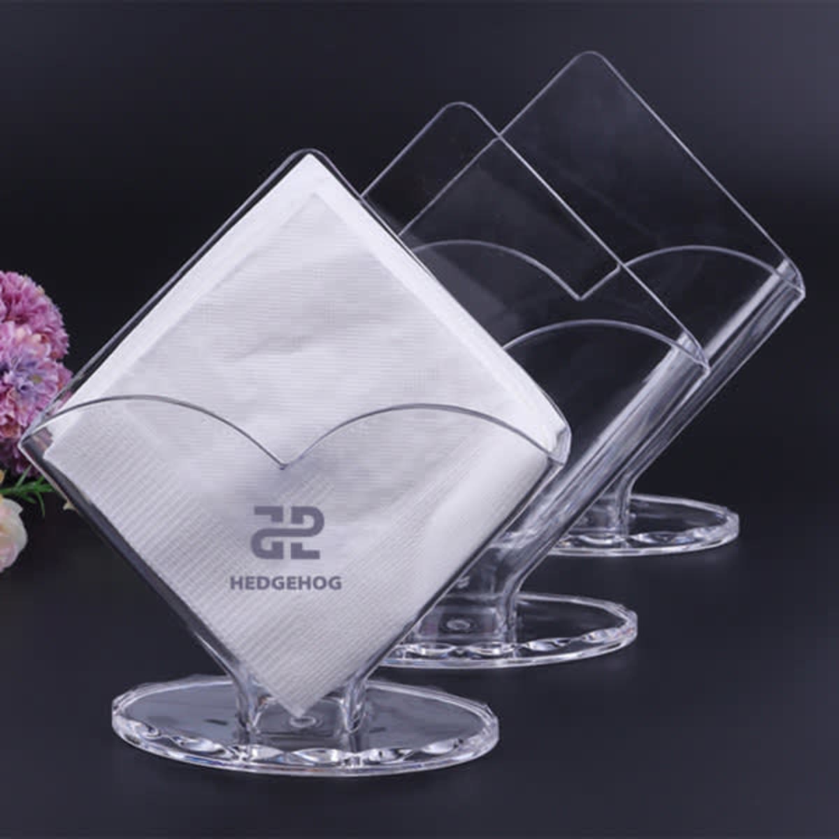 Three clear napkin holders with white napkins, arranged in a row on a black surface.
