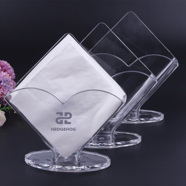 Clear napkin holders with white napkins, arranged in a row on a dark surface.