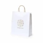 White paper shopping bag with handles and a gold floral emblem design on the front.