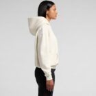 Person in a cream-colored hoodie and black pants standing in profile against a plain background.