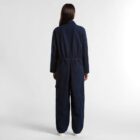 Person standing facing away, wearing a dark blue jumpsuit and white shoes on a plain background.