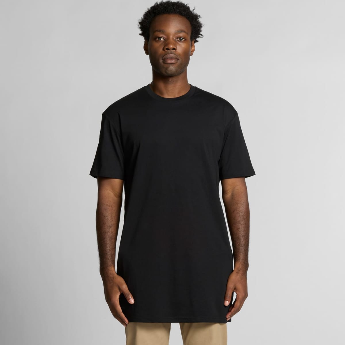 A man wearing a plain black t-shirt and beige pants stands against a light gray background.