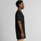 Man in a black t-shirt and tan pants standing in profile against a plain background.