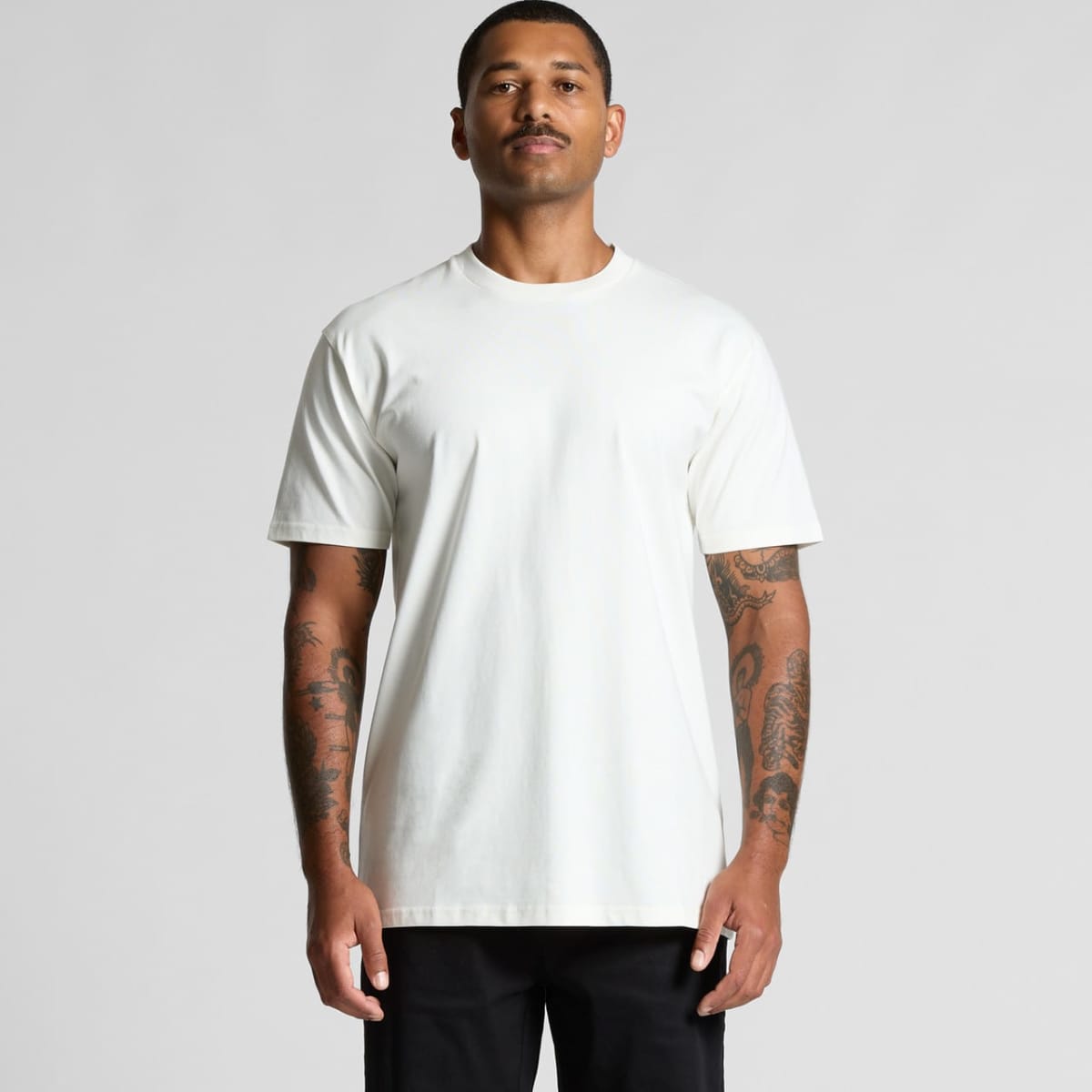 Man with tattoos wearing a plain white t-shirt and black pants, standing against a gray background.