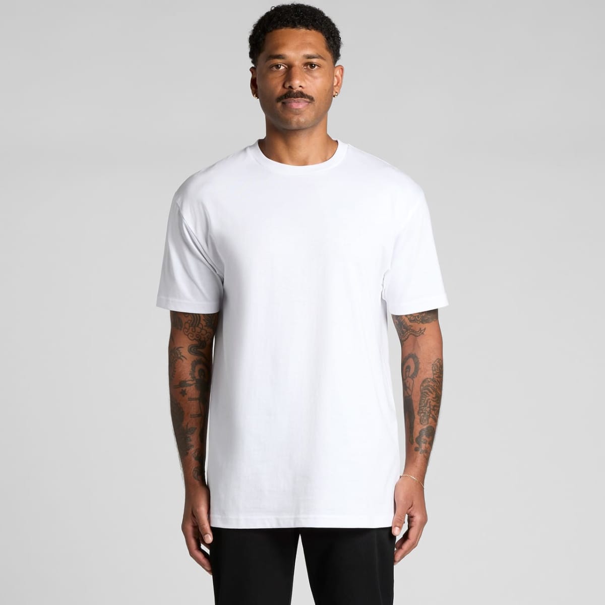 Man with tattoos wears a plain white t-shirt and black pants, standing against a gray background.