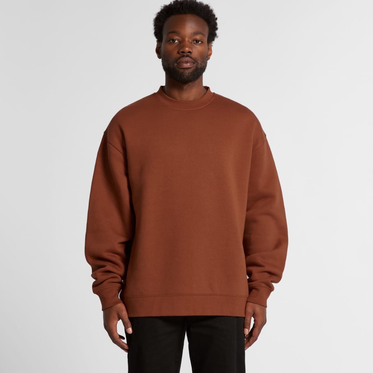 A man wearing a loose brown sweatshirt and black pants stands against a plain background.